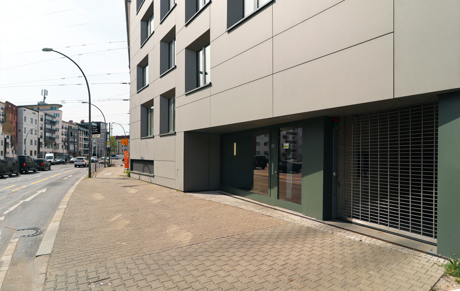 Blick entlang des gepflasterten Gehwegs auf das Erdgeschoss des Gebäudes mit Faserzementfassade, verglastem Bereich und Metallgitter, städtisches Umfeld mit Straßenlaterne.