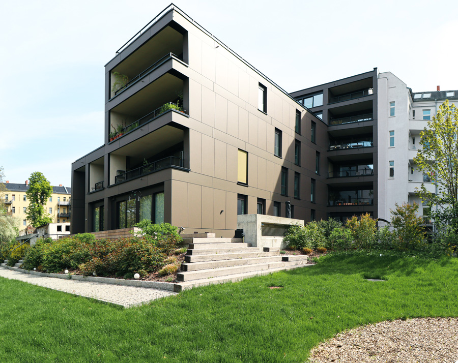 Gartenseitige Fassade des Wohnhauses mit anthrazitfarbener Platten- und Balkonfront, außen liegender Treppe und Rasenfläche bei Sonnenschein, rechts ein angrenzender Altbau.