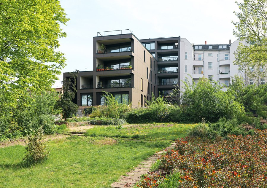 Rückseite des Mehrfamilienhauses mit dunkler Fassade und gestapelten Balkonen, gesehen über eine begrünte Gartenanlage mit Sträuchern und Weg.