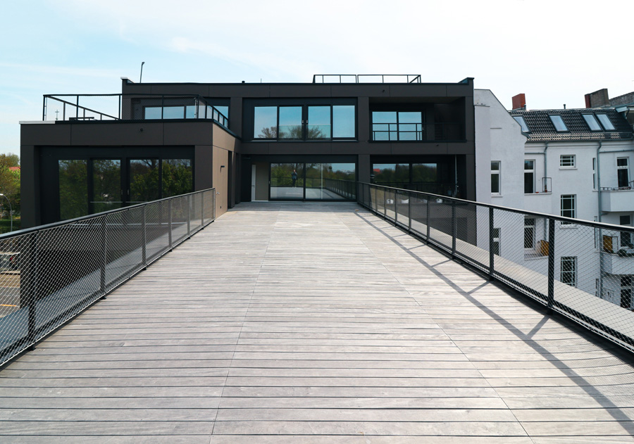 Große Dachterrasse mit Holzdielenbelag und Drahtnetzgeländer vor dem zurückgesetzten Penthouse-Geschoss mit dunkler Fassade; rechts ein Nachbar-Altbau unter blauem Himmel.