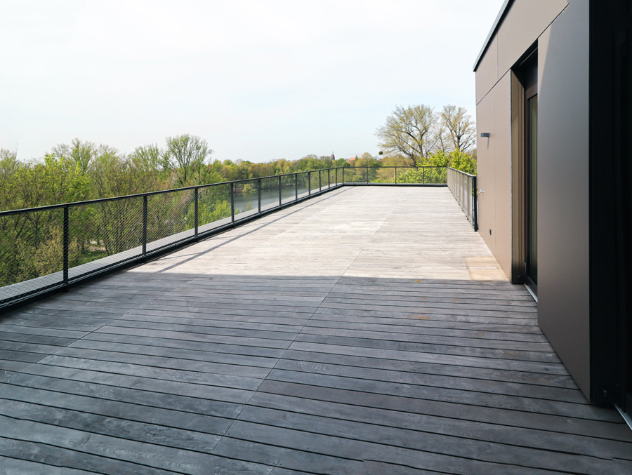 Weitläufige Dachterrasse mit Holzdeck und Drahtnetzgeländer, Ausblick über Baumkronen auf ein Gewässer.