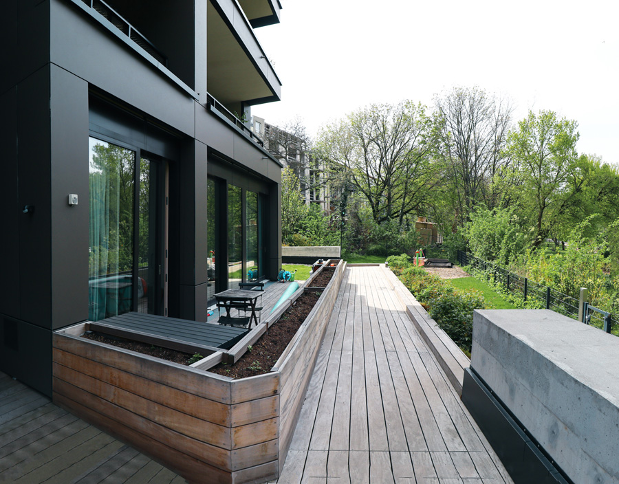 Umlaufende Terrasse an der Gebäuderückseite mit Holzdielen und hölzernen Pflanzkübeln, Blick in den begrünten Garten.