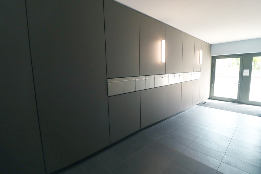 Eingangsbereich mit dunkel verkleideter Wand, Briefkastenanlage und verglaster Hauseingangstür, grauer Fliesenboden.