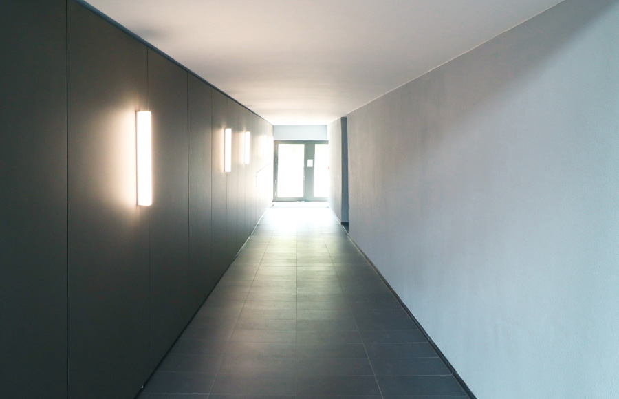 Innenflur mit dunkel verkleideter Wand, Wandleuchten und grauem Fliesenboden, am Ende eine verglaste Tür mit Tageslicht.