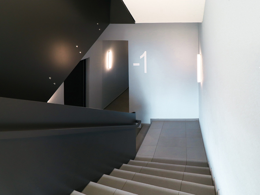 Treppenhaus mit grau gefliesten Stufen, dunklem Handlauf und Wandleuchten, Geschossbeschriftung an der Wand.