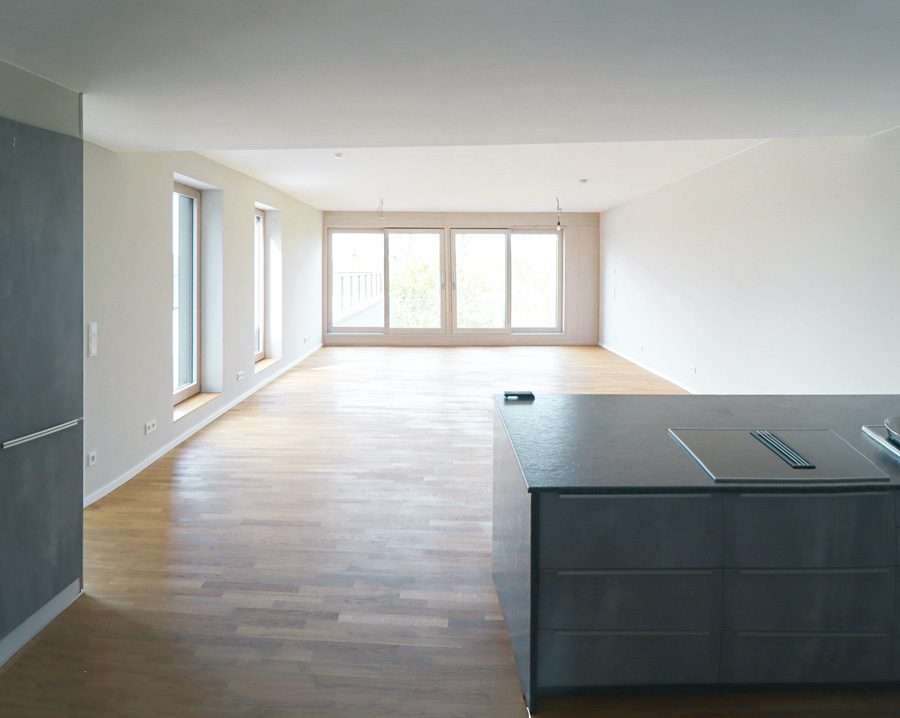 Offener Wohn- und Küchenbereich einer leeren Wohnung mit Parkettboden, Kochinsel mit dunkler Steinarbeitsplatte und Fensterfront zum Balkon.