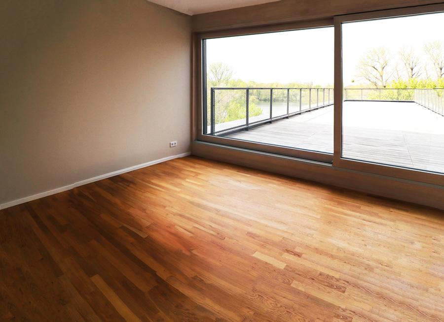 Leerer Wohnraum mit Parkett, taupefarbenen Wänden und raumhoher Eckverglasung mit Blick auf Terrasse und Gewässer.