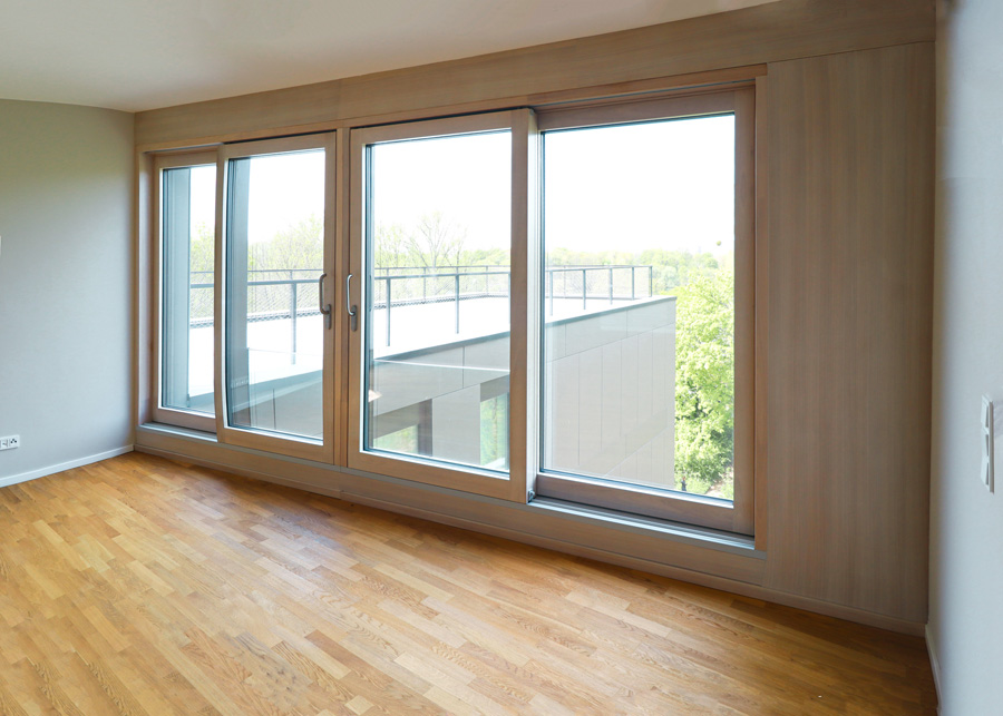 Raum mit Parkettboden und großer holzgerahmter Schiebefensterfront, Blick auf eine Terrasse und Bäume.
