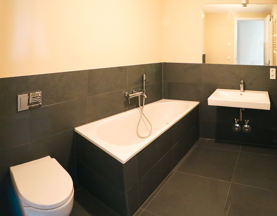 Badezimmer mit Badewanne, Wand-WC und Waschbecken, dunkle Wand- und anthrazitfarbene Bodenfliesen.