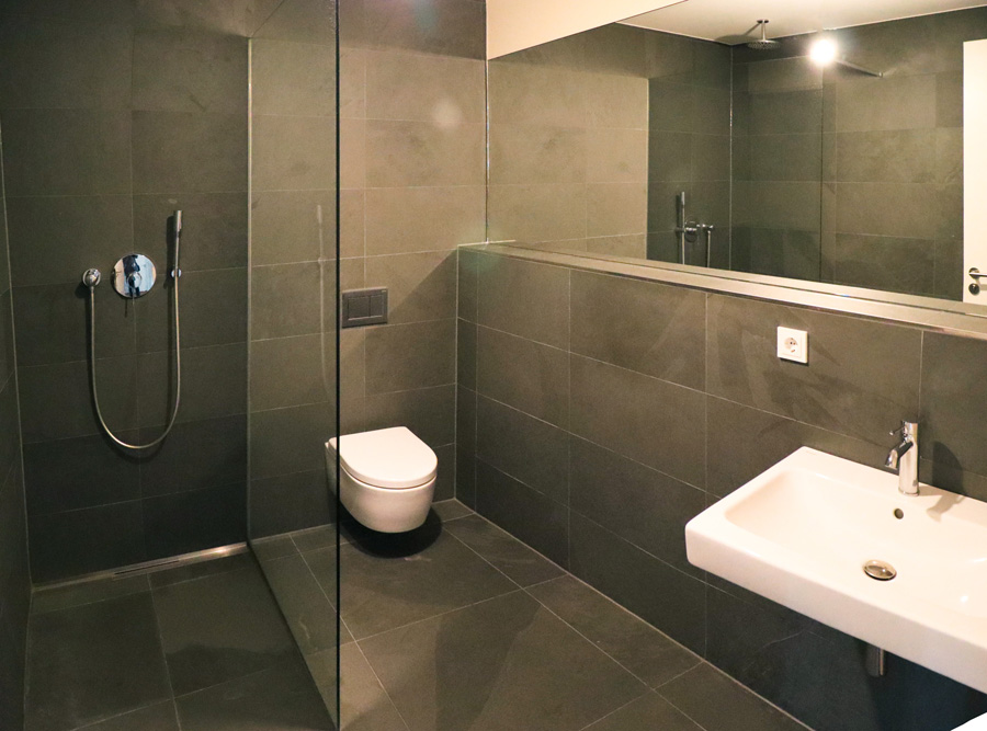 Bad mit bodengleicher Dusche hinter Glastrennwand, Wand-WC, Waschbecken und breitem Wandspiegel, durchgehend dunkel gefliest.