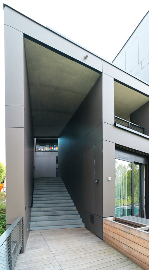 Architekturdetail: überdachte Außentreppe zwischen zwei Baukörpern mit anthrazitfarbener Plattenverkleidung, Holzdeck im Vordergrund.
