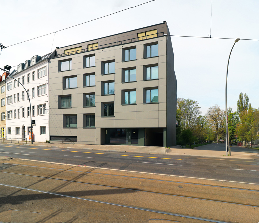 Straßenansicht des mehrgeschossigen Mehrfamilienhauses über Eck bei Sonnenschein, mit grauer Faserzement-Plattenfassade und zurückgesetztem Staffelgeschoss, an einer Straße mit Straßenbahngleisen neben einem hellen Altbau.
