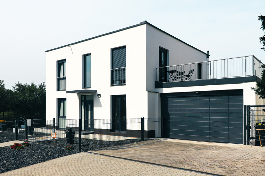 Weißes Flachdachhaus mit integrierter Garage mit anthrazitfarbenem Sektionaltor, Dachterrasse mit Metallgeländer und gepflasterter Zufahrt hinter einem Metallzaun.