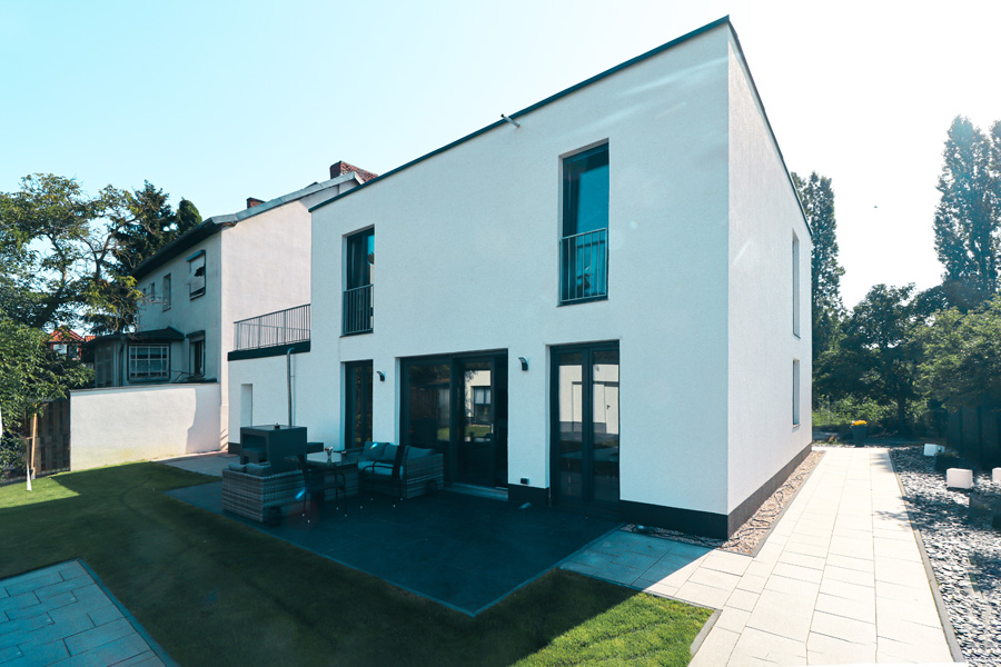 Gartenseitige Rückfassade des weißen Flachdachhauses mit ebenerdiger Terrasse, dunklen Loungemöbeln und umlaufender Rasenfläche neben dem älteren Nachbargebäude.