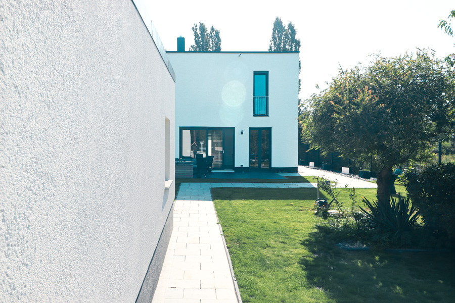 Seitliche Perspektive entlang der weißen Putzfassade mit gepflastertem Gartenweg, Rasen, Baum und rückwärtiger Terrasse mit bodentiefen Fenstern.