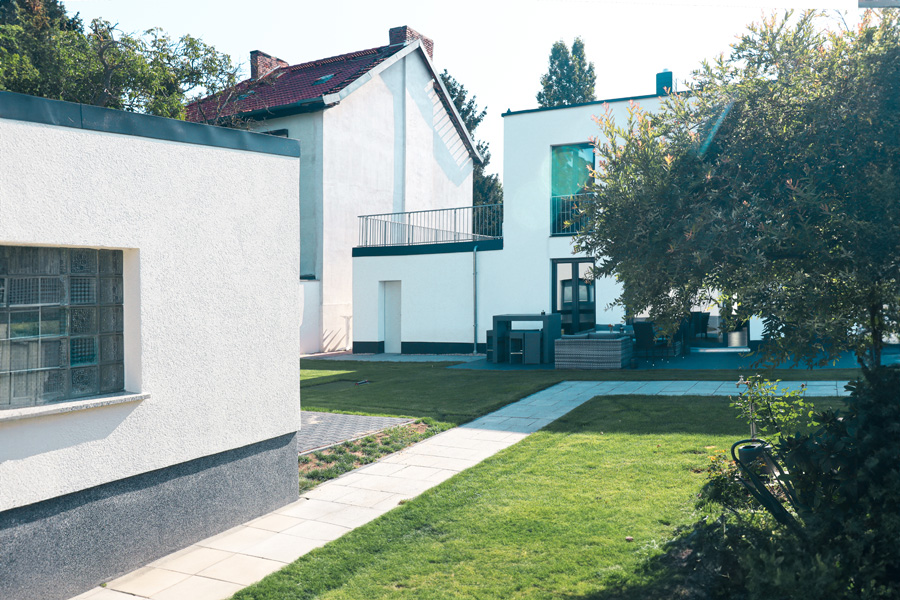 Gartenansicht mit weißem Nebengebäude mit Glasbaustein-Fenster im Vordergrund und dem Haupthaus mit Dachterrasse im Hintergrund, dazwischen Rasen und Wegeplatten.