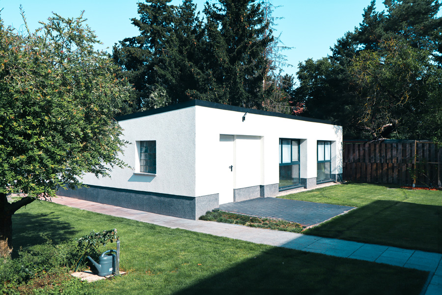 Freistehendes weißes Nebengebäude mit Flachdach und anthrazitfarbenem Sockel im begrünten Garten, umgeben von Bäumen, Rasen und einem Holzzaun.