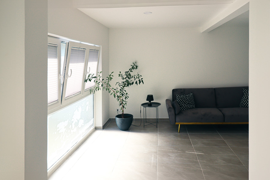 Heller Wohnraum mit grauen großformatigen Bodenfliesen, dunkelgrauem Sofa, Zimmerpflanze und bodentiefem Fenster mit Plissee.