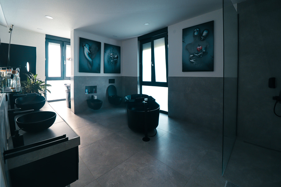 Badezimmer mit großformatigen grauen Fliesen, freistehender schwarzer Badewanne, zwei schwarzen Aufsatzwaschbecken, wandhängendem WC und ebenerdiger Dusche.