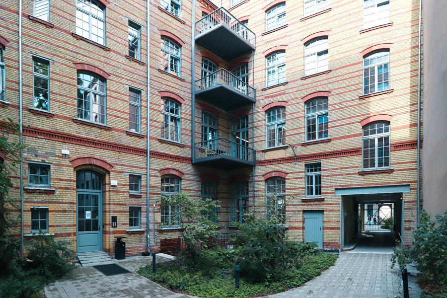 Restaurierter Innenhof mit gelb-roter Klinkerfassade, mittigem Stahl-Balkonturm, blauer Hauseingangstür und neu gepflastertem, begrüntem Hof.