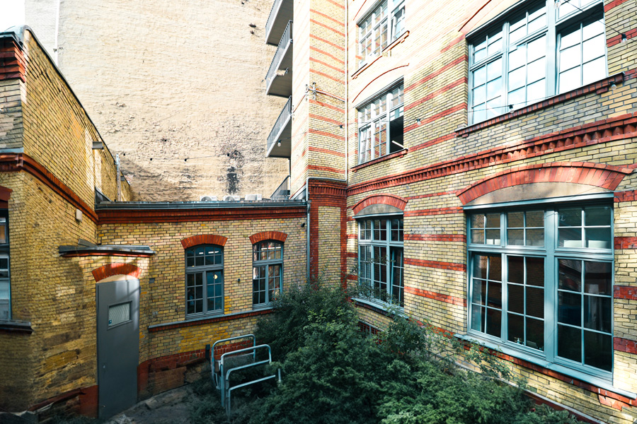Innenhof-Ecke mit gelb-roter Klinkerfassade und großen Sprossenfenstern, dahinter eine hohe verputzte Brandwand.