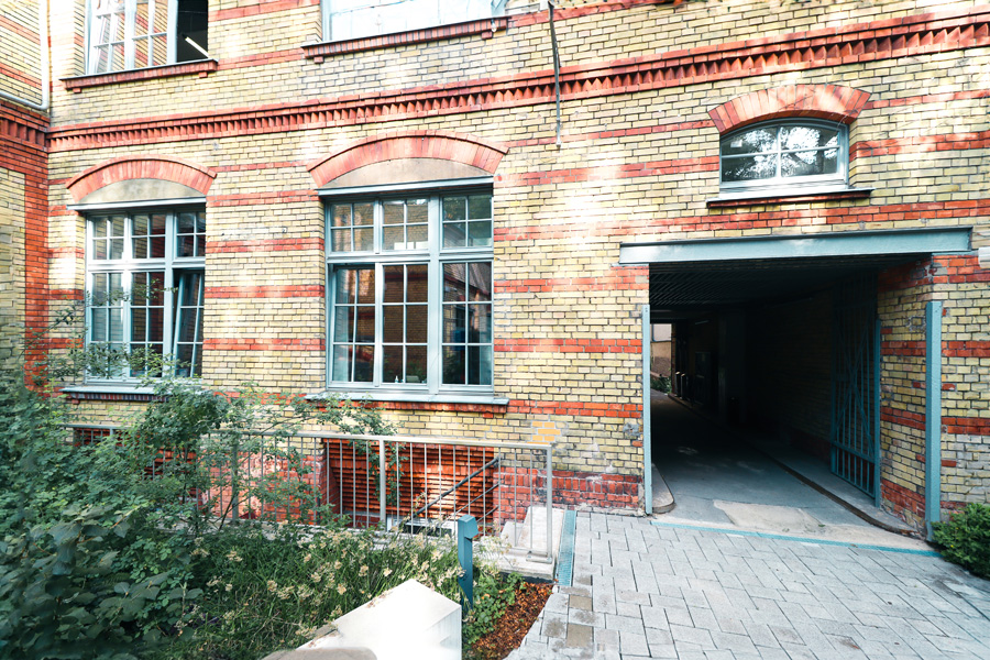 Klinkerfassade im Hof mit Sprossenfenstern über roten Segmentbögen und offener Toreinfahrt, davor Beetbepflanzung.