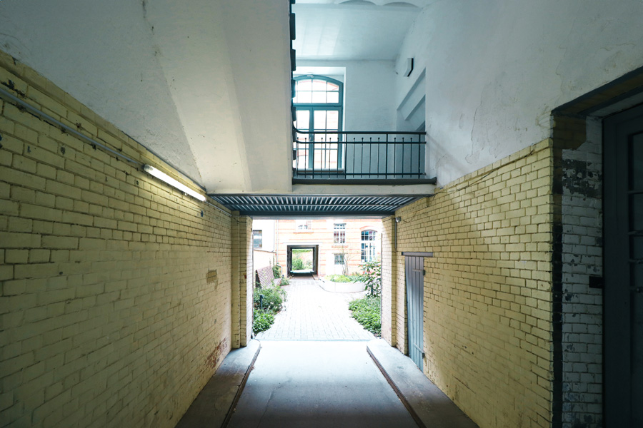 Überdachte Tordurchfahrt mit gelb glasierten Ziegelwänden und sichtbarer Stahlkonstruktion, mit Blick auf den begrünten Innenhof.