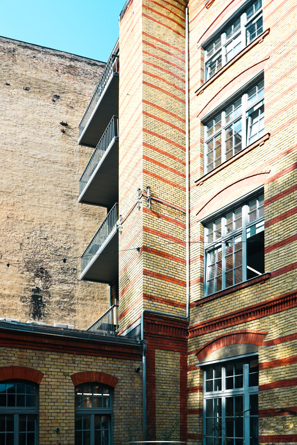 Detailaufnahme des vorgesetzten Stahl-Balkonturms an der bandartig rot-gelben Klinkerfassade vor einer verputzten Brandwand.