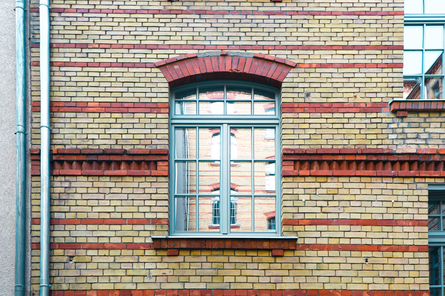 Detail eines Sprossenfensters mit rotem Klinker-Segmentbogen und metallenem Regenfallrohr in der gelb-roten Klinkerfassade.