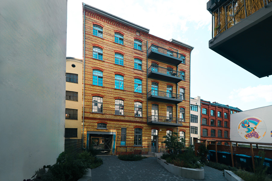 Gesamtansicht des fünfgeschossigen Mehrfamilienhauses mit gelb-roter Klinkerfassade, angebautem Stahl-Balkonturm und Toreinfahrt im Erdgeschoss, davor ein gepflasterter Vorplatz mit Bepflanzung.