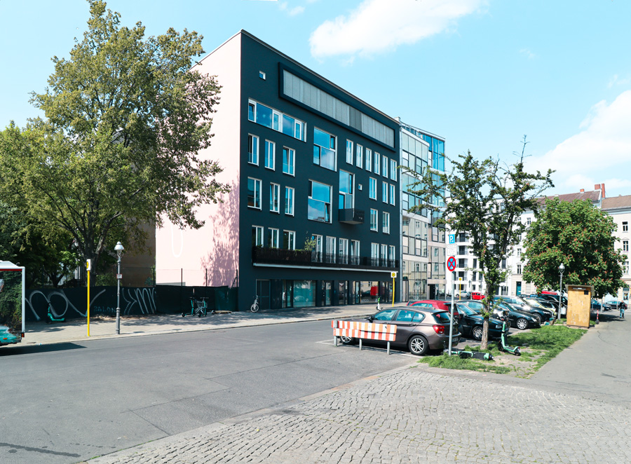 Gebäudeecke an einer Berliner Straßenkreuzung mit Kopfsteinpflaster: dunkle anthrazit-grüne Straßenfassade und angrenzende rosafarbene Giebelwand, davor parkende Autos und Straßenbäume.