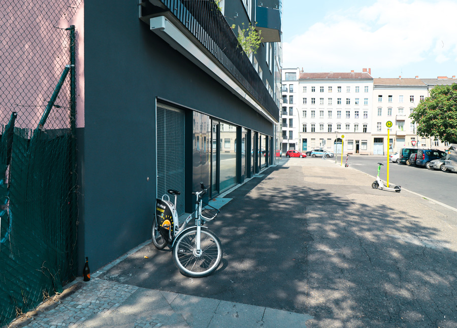 Erdgeschosszone der dunklen Fassade auf Straßenniveau mit Schaufenstern und angelehntem Leihfahrrad, gegenüber Berliner Altbauten.