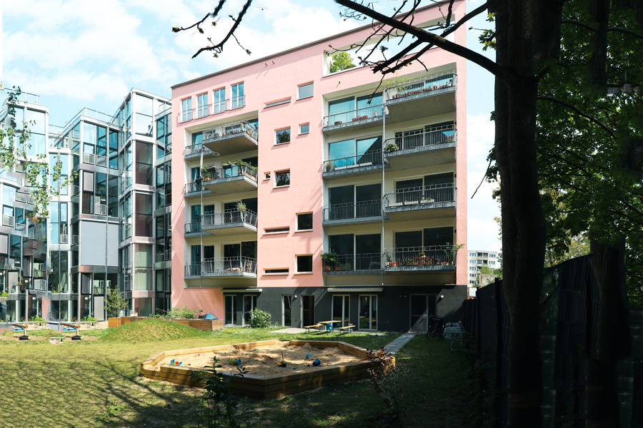 Hofseitige rosafarbene Fassade mit umlaufenden Balkonen, davor begrünter Innenhof mit hölzernem Sandkasten und Rasenfläche.