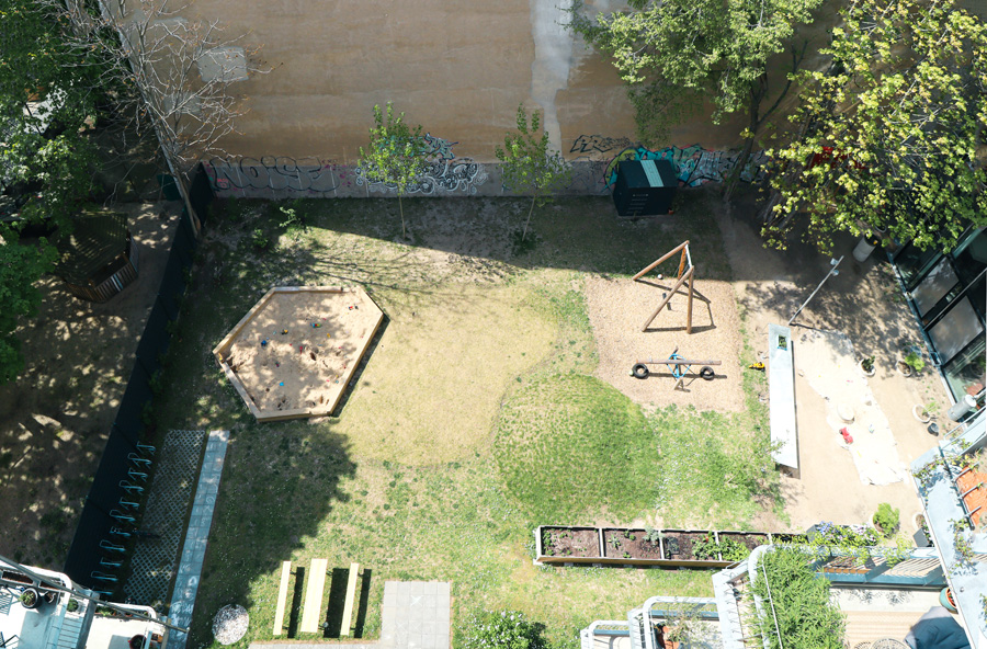 Luftaufnahme des begrünten Innenhofs mit Sandkasten, Spielgeräten, Sitzbänken und Hochbeeten zwischen den Gebäuden.