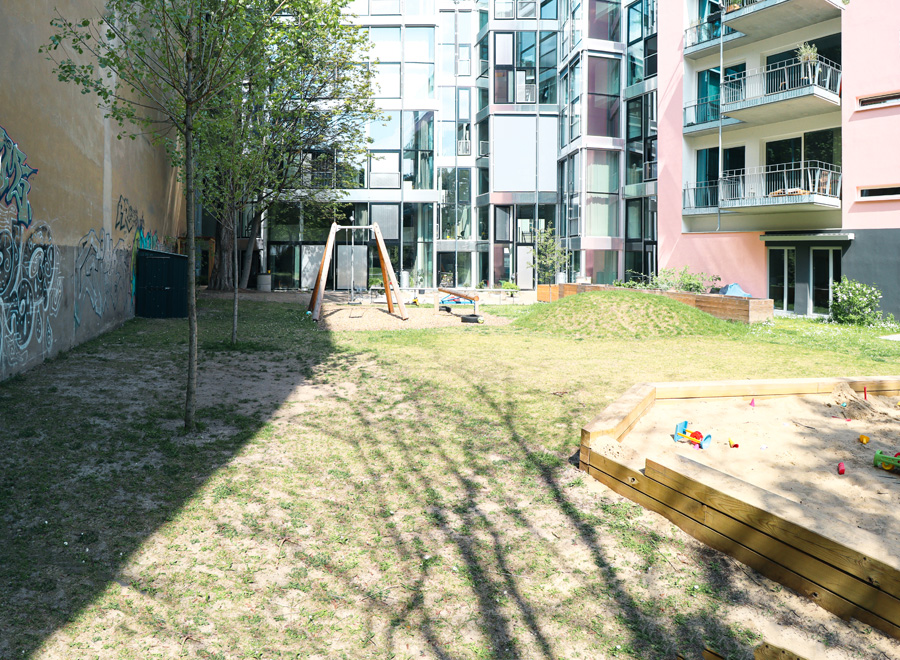 Innenhof mit Schaukelgerüst und Sandkasten auf Rasen, im Hintergrund eine mit Graffiti bedeckte Brandwand und ein verglastes Nachbargebäude.