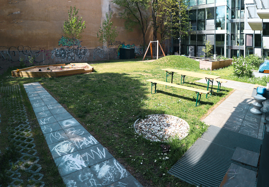 Innenhof mit Sitzbänken, Feuerstelle, aufgemaltem Hüpfspiel auf Gehwegplatten und Sandkasten vor einer graffitibedeckten Brandwand.