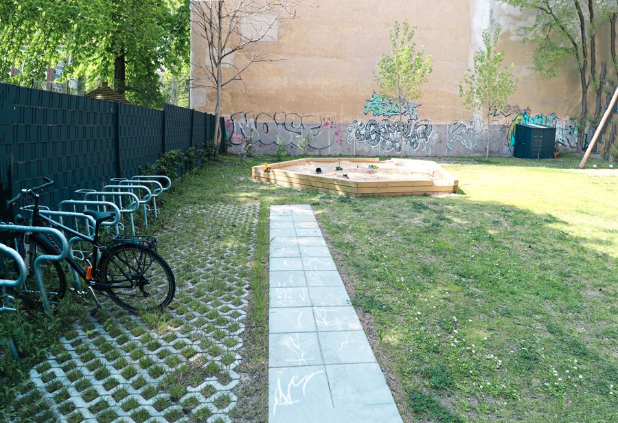 Hofbereich mit überdachten Fahrradständern, Plattenweg und Sandkasten vor einer mit Graffiti bedeckten Brandwand.