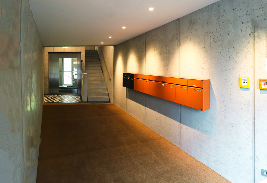Hauseingangshalle mit Sichtbetonwänden, orangefarbener Briefkastenanlage, Treppenaufgang und gemustertem Bodenbelag.