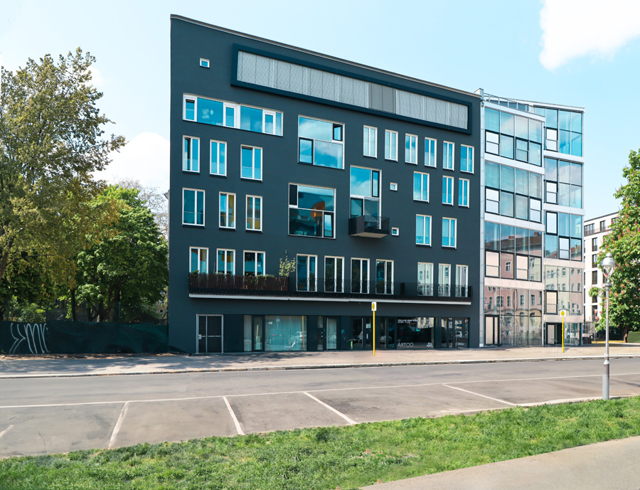 Straßenfassade des mehrgeschossigen Wohn- und Geschäftshauses mit dunkler anthrazit-grüner WDVS-Fassade, gereihten Fenstern, kleinen Balkonen und verglasten Ladenzonen im Erdgeschoss.