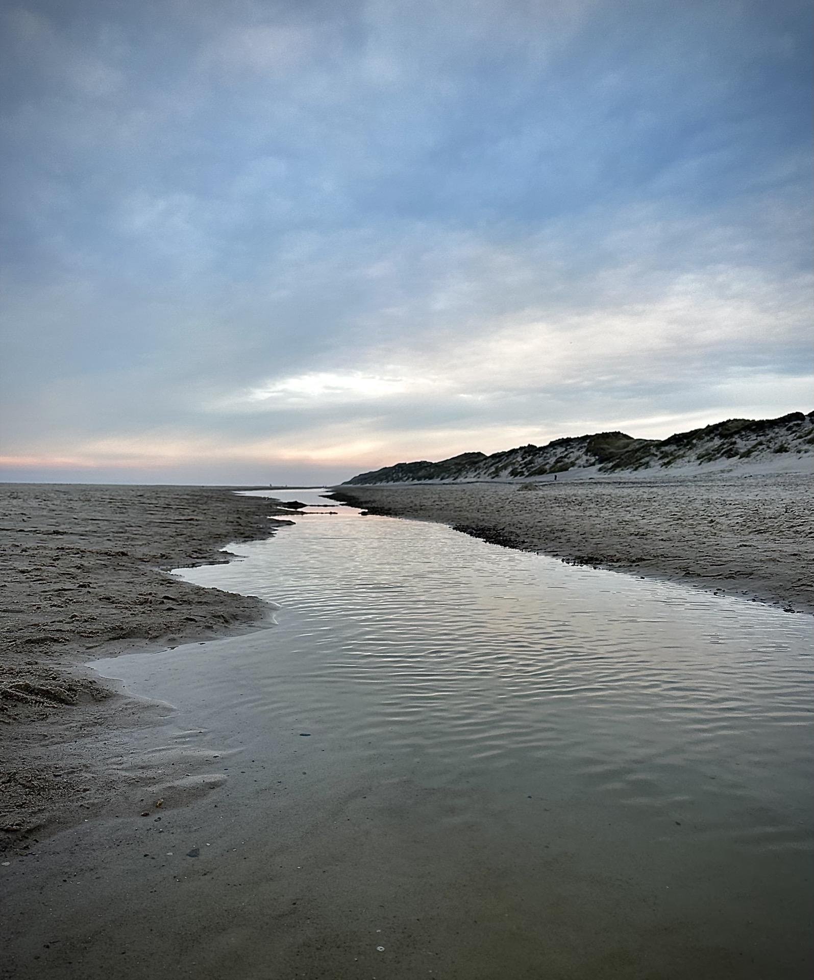 This image captures a serene beach scene with a gentle waterway meandering through the sand. On either side, there are sandy dunes lightly covered in grasses and shrubs, typical of coastal landscapes. The sky has a soft gradient of blues and faint pink, possibly indicating late afternoon or early evening. The tranquility of the scene, along with the natural curvature of the water's path, creates a peaceful, picturesque vista.