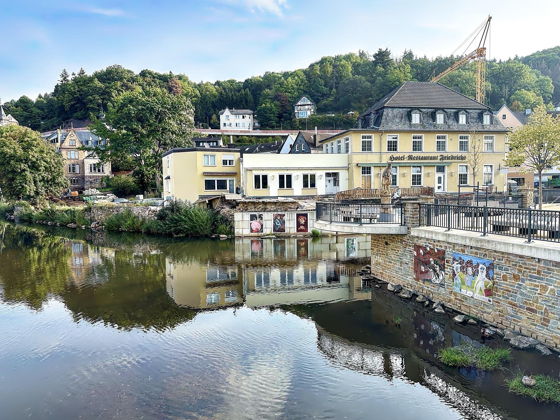 This image depicts a scenic riverside view featuring a mix of commercial and residential buildings. The central structure is labeled as "Hotel-Restaurant Rheinblick," suggesting it's a hotel with a restaurant that probably offers views of the river. The environment is peaceful with lush greenery in the background, including trees and hillside homes that add to the charming setting. There's a calm river in the foreground that beautifully reflects the buildings and the sky, enhancing the serene and picturesque quality of the scene. The architecture and the layout indicate a European setting, possibly in a small town known for its hospitality and beautiful landscapes.