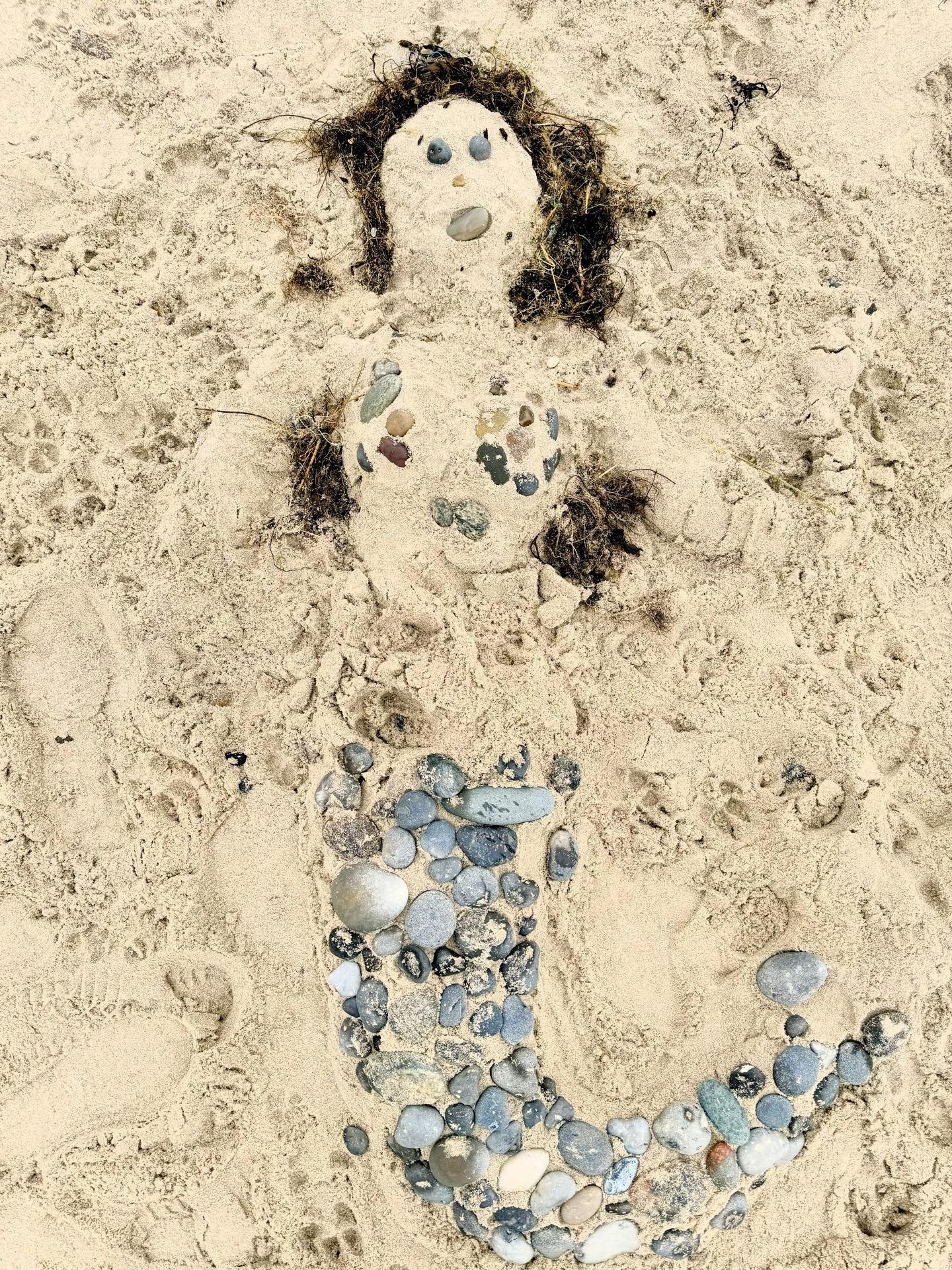The image shows a creative representation of a mermaid made from various materials on a sandy beach. The mermaid figure is constructed with a head, body, and tail, all made from sand. The face features eyes, a nose, and a mouth created using small pebbles, and seaweed is used for the hair. The torso and tail of the mermaid are adorned with pebbles in various shapes and colors, arranged to enhance the shape of the body and the swirling tail. This artistic beach creation uses natural materials found in the environment, displaying a playful use of beach elements to craft a scene.