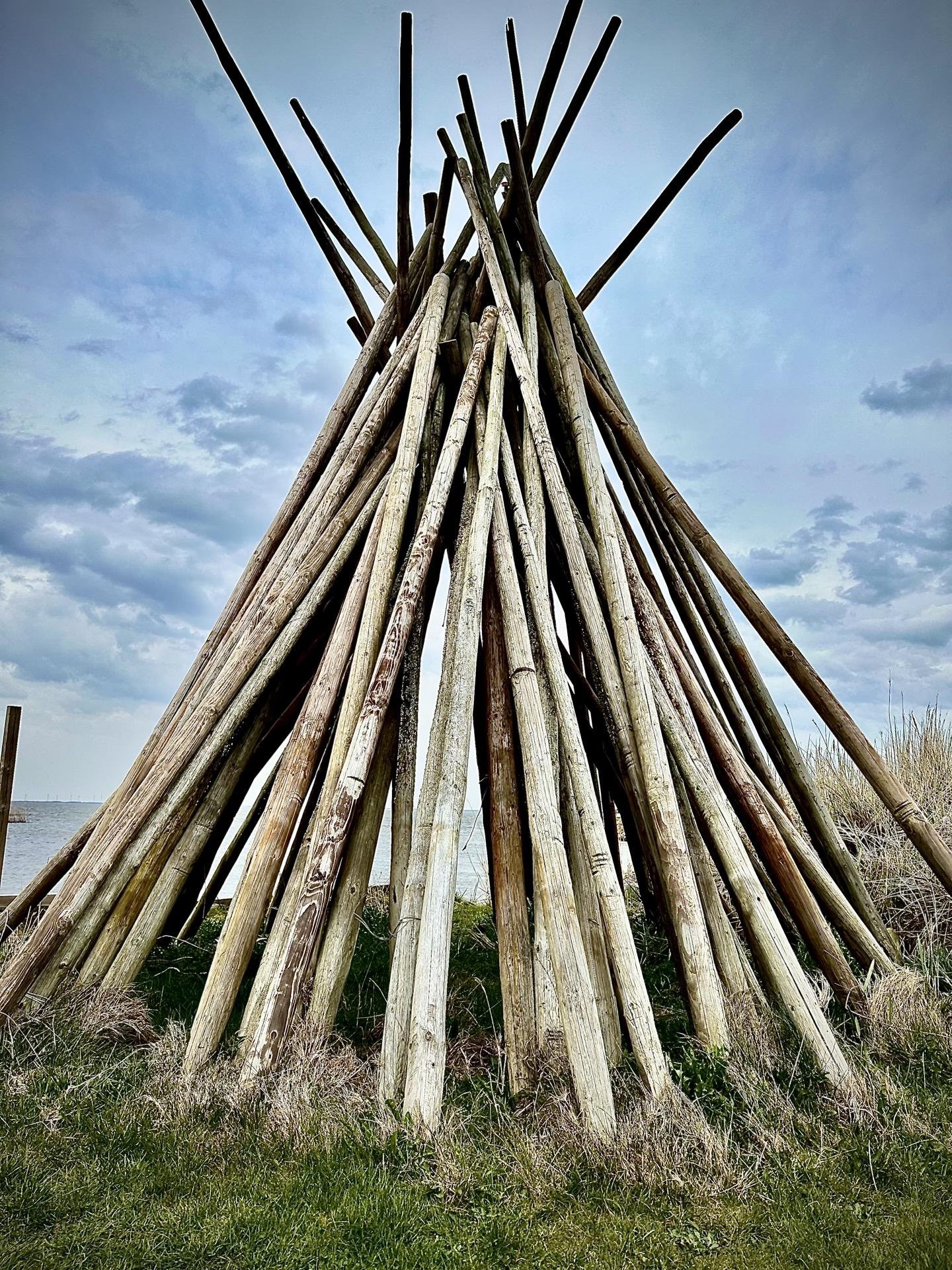 This image features a large, cone-shaped structure made from many long wooden poles or logs arranged leaning together at the top, reminiscent of a teepee construction. It's located in a natural setting with visible grass in the foreground and a body of water and overcast sky in the background. The structure likely serves a decorative or functional role in this outdoor setting, possibly used for shelter or as a visual accent in the landscape.