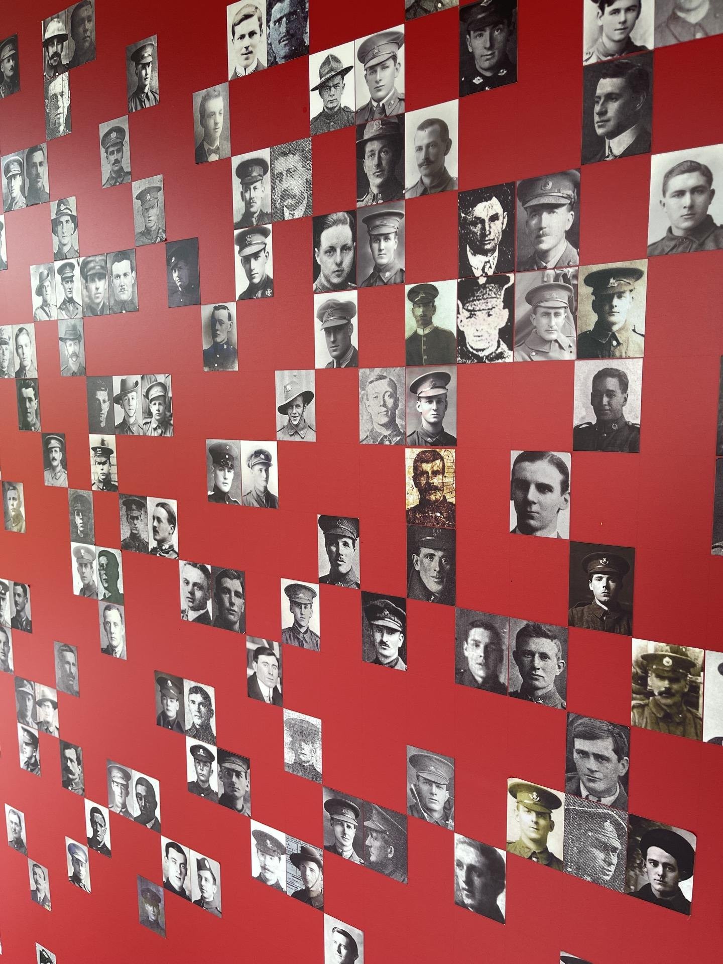 This image shows a collection of black and white photographs arranged on a red wall. Each photograph portrays an individual, predominantly males, most likely soldiers or military personnel given that several are wearing uniforms featuring caps or hats typical of military attire. The style of uniforms and the photographic quality suggest that these images could be historical, possibly from the early to mid-20th century. The red background creates a striking contrast with the black and white photos, emphasizing each individual portrait. This display could be part of a historical exhibit, memorial, or a tribute to military personnel from a specific period.