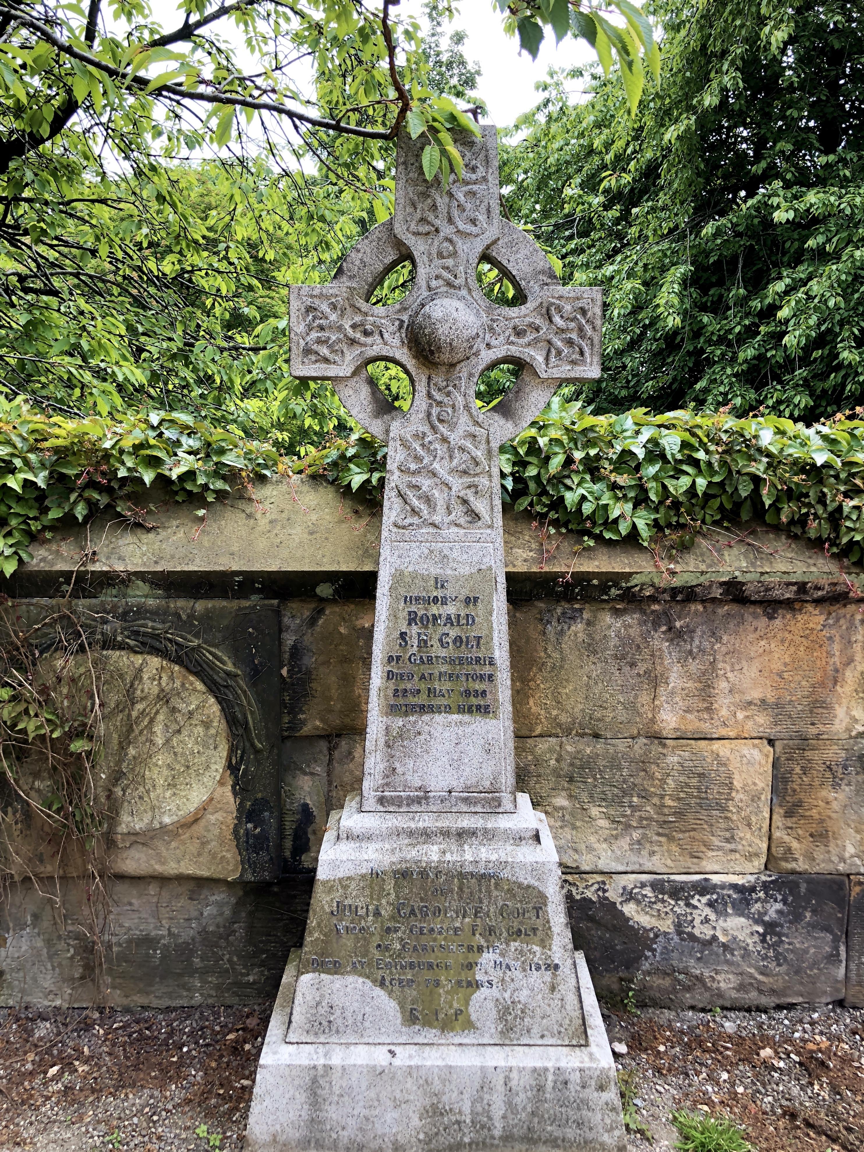 This image shows an ornate stone Celtic cross gravestone. The cross features intricate knotwork designs, a common motif in Celtic art, emphasizing circular forms and interlacing patterns. The inscription on the gravestone commemorates individuals named Ronald S.F. Colt and Jean Cardin F.H. Colt. It mentions that Ronald S.F. Colt died on 22 May 1936 and was interred at the location of the gravestone. Jean died on 10 May 1938 at the age of 71 years. The stone cross stands before a wall covered with some ivy and is surrounded by verdant vegetation, likely located in a serene cemetery setting.