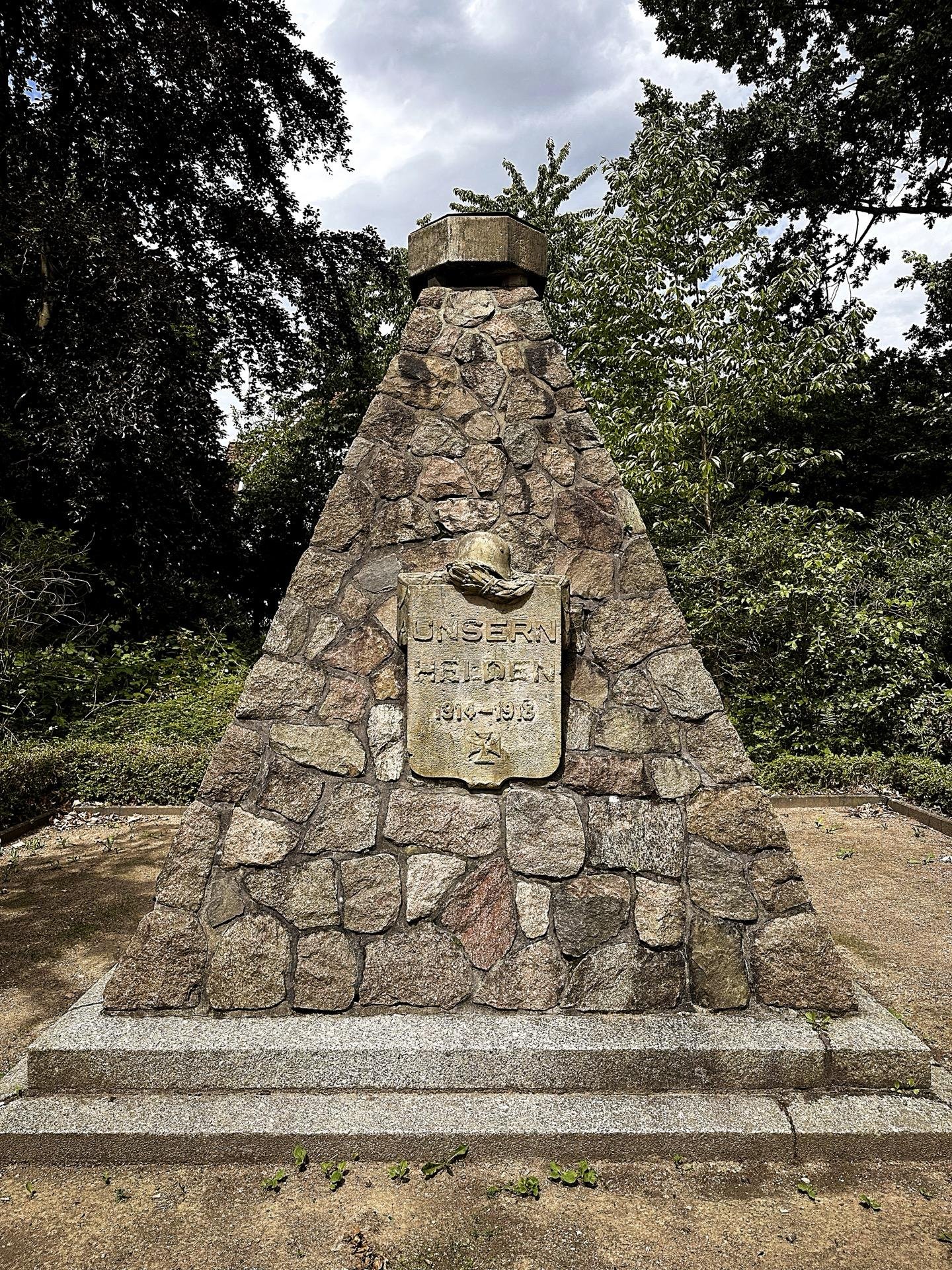 The image shows a memorial built from stones, structured in a triangular or pyramidal shape with a capstone on the top. There is a plaque attached to the monument which reads "UNSERN HELDEN 1914-1918", likely a tribute to fallen soldiers from World War I, indicating this is a war memorial dedicated to "Our Heroes" from the years 1914 to 1918. The monument is set in a serene location with trees and greenery surrounding it.