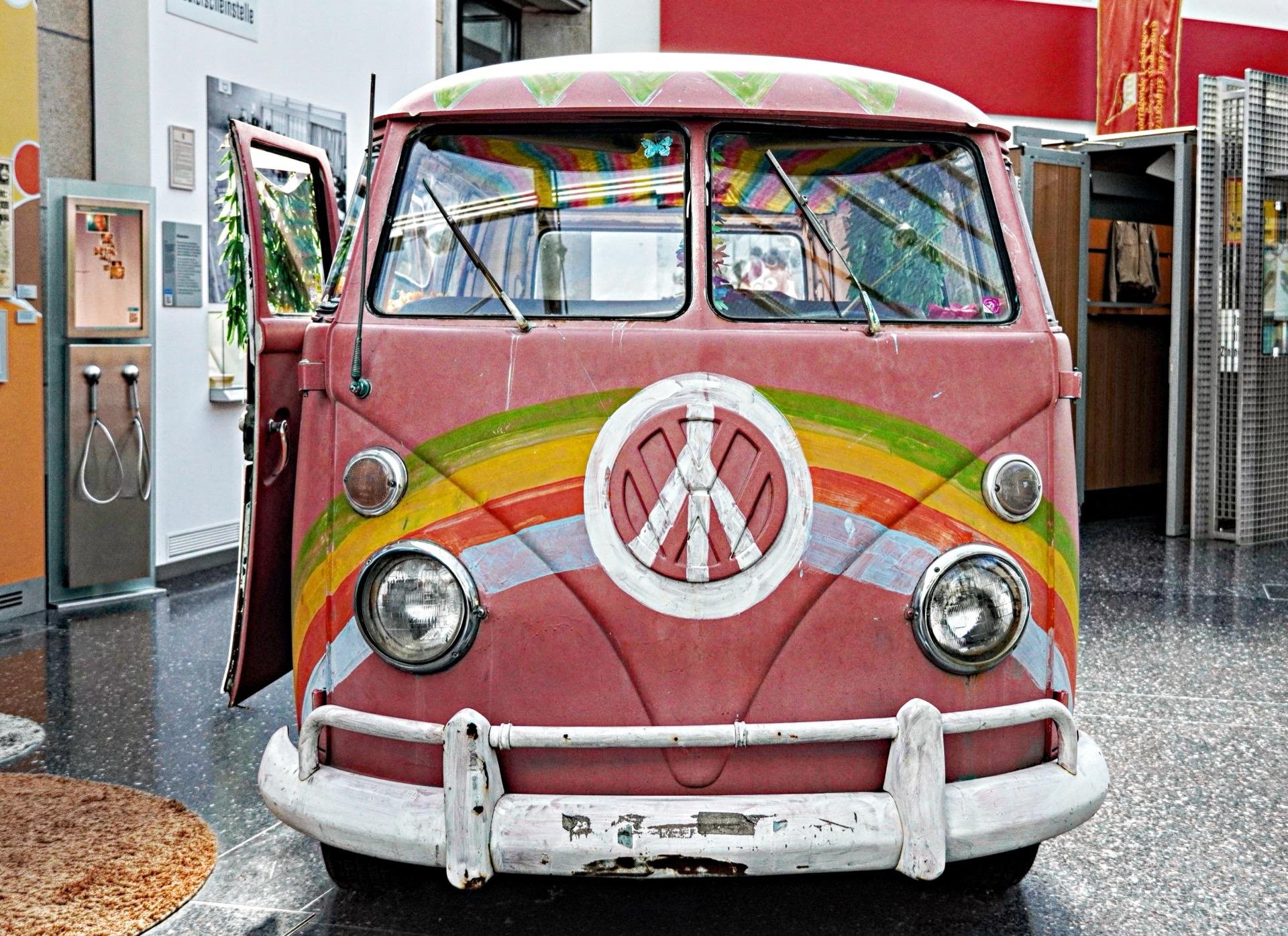 This image features a vintage Volkswagen Type 2 van, often referred to as a VW Bus or Kombi. The van is painted in a colorful, psychedelic pattern that includes swirls and rainbow colors, prominently displaying the Peace symbol on the front. The van's front doors are open, and it appears to be on display indoors, perhaps as part of an exhibition or at a museum, evident from its placement on a polished floor and the surrounding informational displays. The overall vibe of the van suggests it may be representative of 1960s counterculture, often associated with the hippie movement.