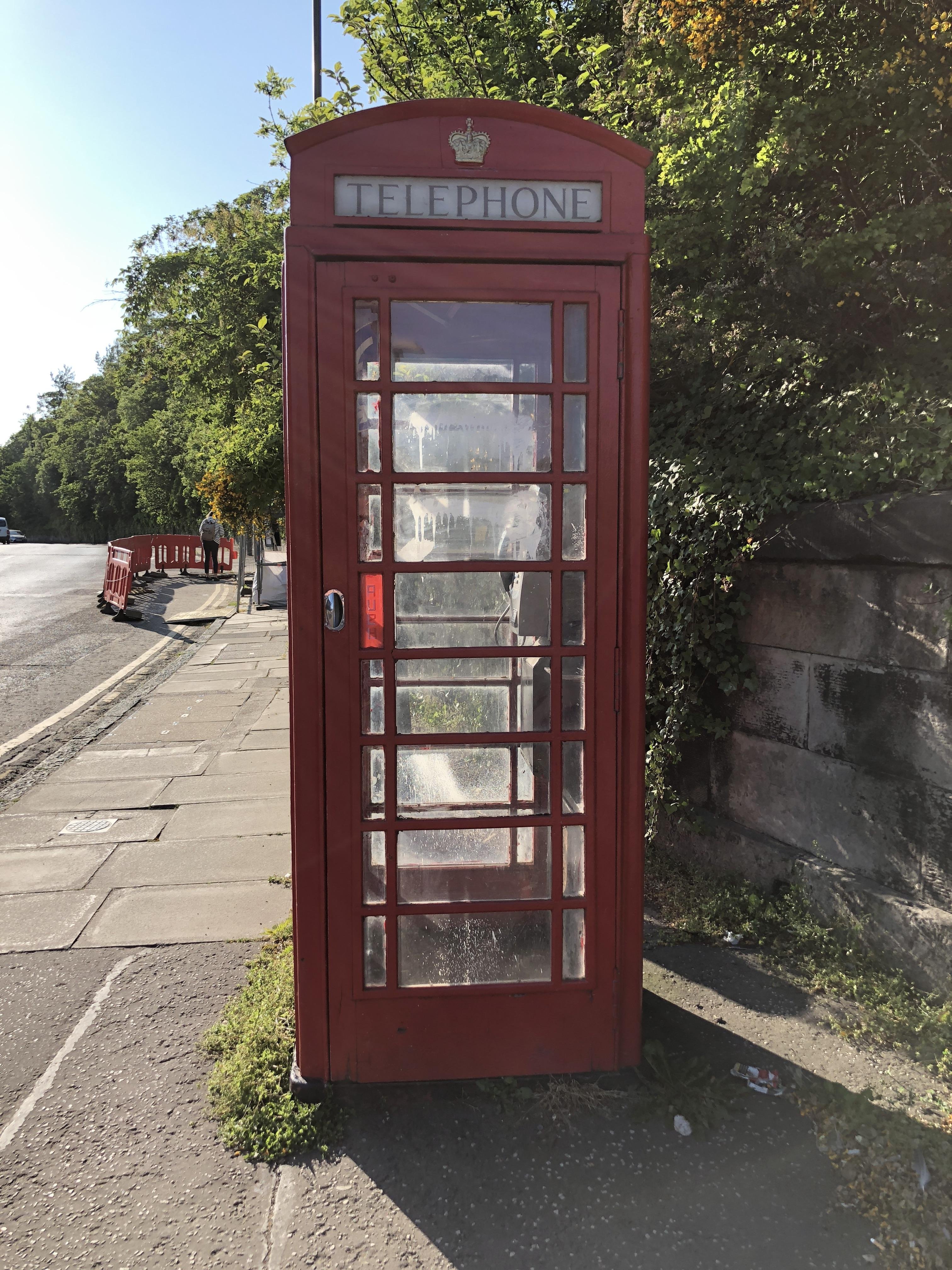 The image shows a traditional British red telephone box located on a sidewalk next to a road. The booth is constructed with glass panels on each side and a solid door, displaying the customary design of these iconic phone booths. Above the door, there is a sign that reads "TELEPHONE" under a crown symbol, indicating its official use as a public telephone facility. The telephone box is situated beside a leafy green tree and appears to be in a typically urban setting with a paved sidewalk and a partial view of the street in the background. The sunlight illuminates the scene, creating a bright and clear day.