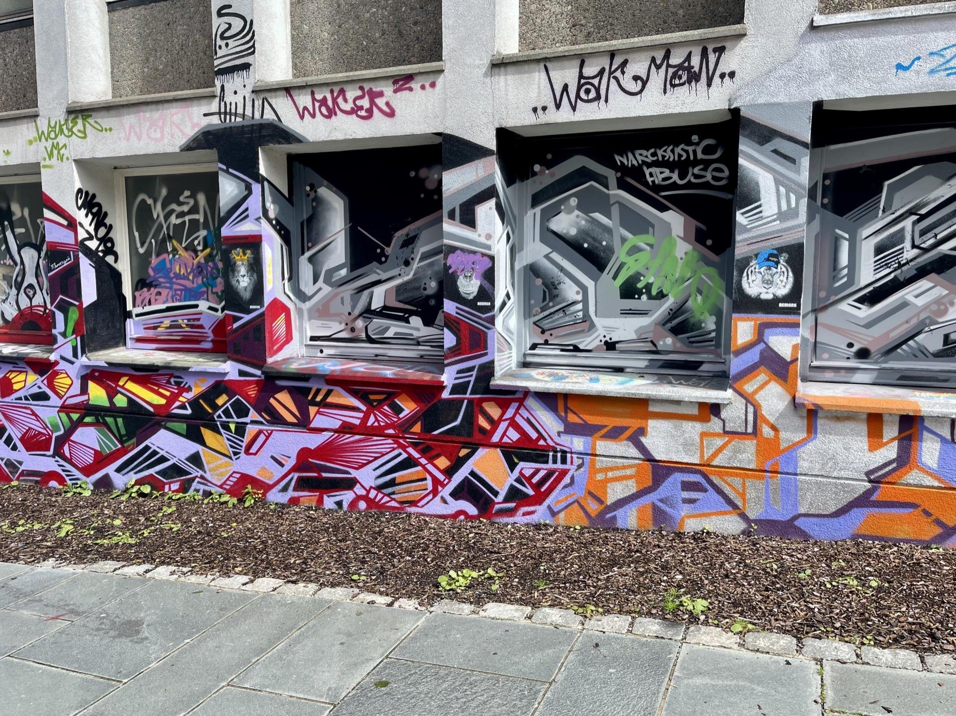 This image shows a vibrant graffiti-covered wall. The graffiti features a variety of styles and colors including 3D-style cubic structures in silver and black tones, and a colorful abstract pattern in red, orange, and purple hues along the lower portion of the wall. There are also some tags and phrases written over the wall, one noticeable phrase being "narcissistic abuse" written in one of the graffiti pieces. The setting appears to be an urban environment, judging from the concrete building and the pavement visible in the picture.