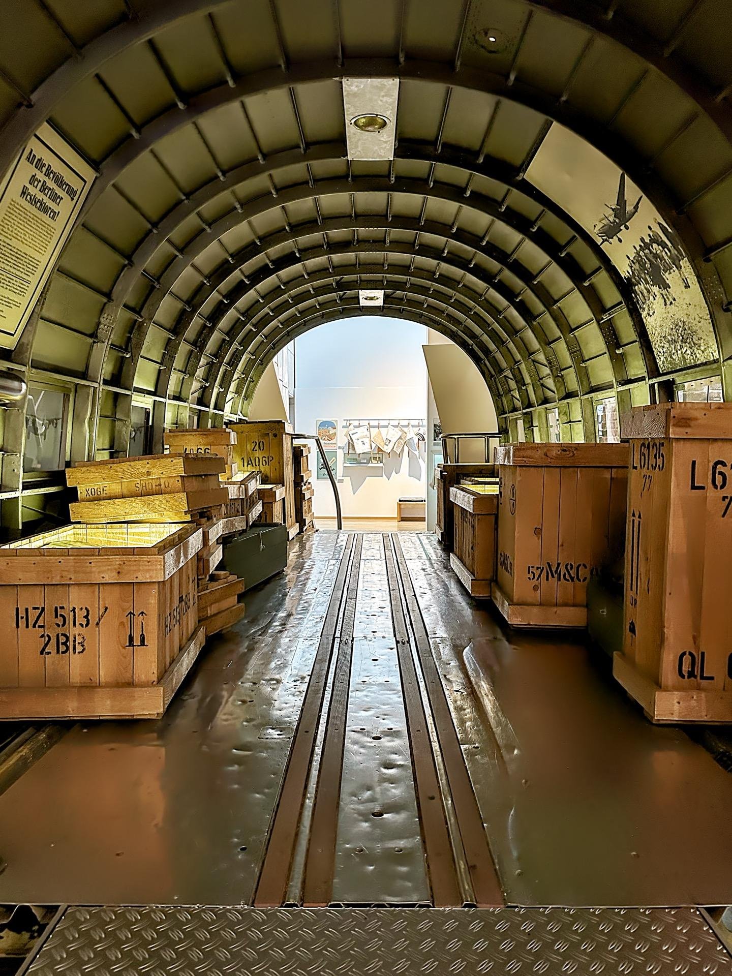 This image shows the interior of a historical military transport aircraft, specifically the cargo hold area. The curved metal ribs of the aircraft's frame create an arched ceiling over the cargo area. There are wooden crates and boxes securely placed within the cargo hold, likely representing supplies such as ammunition, equipment, or other military necessities based on the markings on the crates (like "20 AGB").

The floor of the cargo hold is metal, featuring tracks and tie-downs for securing cargo during flight. Several signs and informational panels are visible, suggesting that this image might have been taken in a museum setting designed to educate visitors about military history and aircraft operations. The inclusion of the historical photograph on the side of the interior further emphasizes the educational purpose of this setting.